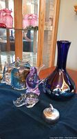 Blue and purple iridescent glass vase and three glass animal figurines on a dark cloth, next to a glass cabinet with pink items inside.