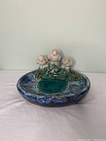 Front view of the ceramic bird bath showing three white ceramic birds sitting on green foliage with a blue basin underneath.