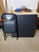 Photo showing black card table and two black folding chairs leaning against the table