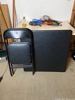 Overall view of the card table folded and two folding chairs leaning against it, placed indoors on a concrete floor.