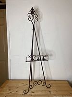Full front view of the metal art easel, showing its height and decorative scrollwork details on the top finial and shelf.