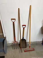 Full view of four garden tools leaning against the wall including a landscape rake, pitch fork, small shovel, and cultivator.