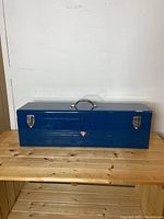 Front view of large blue metal toolbox placed on wooden shelf showing full length, handle, and latches.