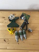 Top down view of lawn care equipment showing the green oscillating sprinkler, black hose nozzle, yellow splitter, three green hose connectors, and metal hose connector on a wooden surface.