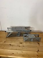 Front and top view of two rectangular galvanized steel live rodent traps on wooden surface against white wall. Larger trap in back, smaller in front.