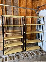 Two metal shelves with black frames and beige shelves, set up in a garage space near a door and wooden walls, including a ladder on floor.
