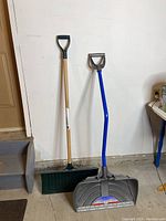 Two snow shovels side by side against a white wall and concrete floor.