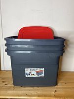Three stacked dark gray storage totes with bright red lids visible inside, front label showing GSC brand and 'Super Heavy Duty Work Box' description with 22 gallon capacity.