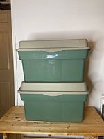 Two stacked Rubbermaid storage totes with green bases and beige hinged lids against a white wall.