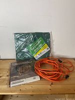 Photo of green tarpaulin, firewood tarpaulin package, and orange extension cord together on wood surface.
