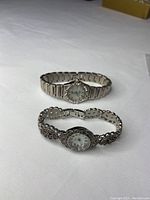 Two silver women's watches on white fabric background, one with a plain metal band and one with decorative band with stones