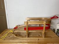 Side view of wooden kids sleigh on wooden table, showing curved backrest and yellow rope attached to front red metal bracket.