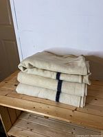 Three folded wool blankets stacked on a wooden surface, showing cream color with a single dark stripe.
