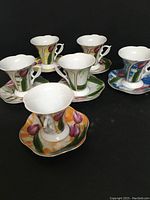Six porcelain espresso cups with flared rims and matching octagonal saucers, each decorated with hand-painted colorful tulip floral designs. Cups have uniquely shaped handles and are shown on a black background.
