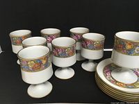 Eight white porcelain dessert cups on pedestal stems with colorful floral band and gold trim, grouped in front of matching plates with floral banding visible.
