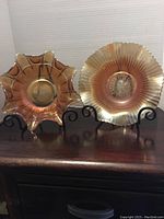 Two carnival glass bowls displayed on black stands. The bowl on the left has a starburst pattern and ruffled edges. The bowl on the right has a more rounded wavy edge and starburst center, both show iridescent golden amber colors.