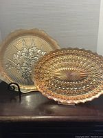 Two carnival glass plates on display, one round with floral pattern and scalloped edge, one oval with diamond pattern and scalloped edge, showing iridescent finish.