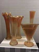 Photo of five carnival glass vases in amber-orange color with iridescent finish, varying heights from 6 to 10 inches, showing ribbed and flared designs.