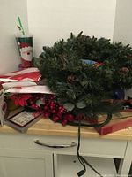 Three artificial wreaths with pinecones and bunches of red berries, boxes likely containing flags or other decorations on a wooden surface.