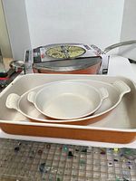 Two cooking pans; a brand new Heritage frying pan with cover on box, and a large orange Le Creuset pan with chips on edges