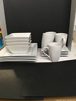 16-piece white ceramic dish set by Canvas with square bowls, square plates, rectangular plates, and mugs on display.