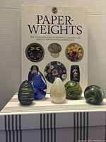 Five decorative glass paperweights arranged in front of a collector's guide book on paperweights, showcasing different colors and shapes.