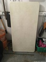 Rectangular folding table with plain light surface, folded flat, leaning against a wall near storage containers