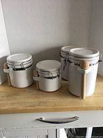 Photo of four white ceramic canisters on wooden surface, showing different sizes and attached wooden spoons