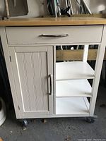 Front view of the kitchen cart showing the drawer with silver handle, cabinet door with vertical grooves, two open shelves, all painted white, and wood top surface.