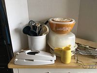Pyrex mixing bowls stacked, plastic containers, rotary egg slicer, yellow pitcher, and kitchen utensils on wooden countertop.