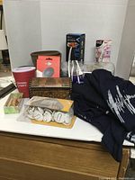 Front view of the miscellaneous lot items arranged on a table, showing an insulated cup, metal box, blue clothing, lights, fan and gloves box