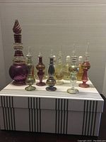 Seven handmade Egyptian perfume bottles arranged on a striped box, showing various colors, shapes, and sizes with tall ornate stoppers.