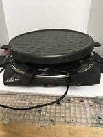 Photo of the black electric raclette grill showing the round textured grilling surface and the black plastic handles of the individual melting trays underneath.