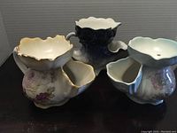 Three porcelain shaving mugs, each with a uniquely shaped leaf-like lather holder attached; floral patterns and gold trim on cream-colored mugs, one blue mug with ventilation holes on top.