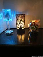 Three touch lamps on dark wooden surface: clear plastic tall lamp lit blue, tree silhouette lamp lit warm yellow, round glass lamp unlit.