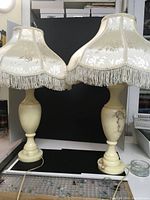 Two alabaster table lamps together showing the bases and cream fabric lampshades with fringe