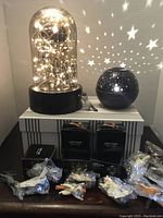 Photo showing two mood lights: a glass dome lamp with warm white fairy lights inside and a star projector night light. Below the lights are five boxes of LED cork string lights and eleven cat-shaped cell phone holders in plastic wrapping.
