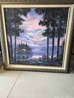 Large framed oil painting showing a lake scene with tall trees in foreground, a cabin near the shore, with mountains and cloudy sky in the background, predominately blue and purple tones.