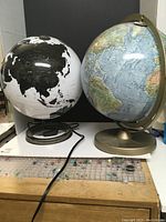 Two world globes shown side by side. The black and white globe is on a metal circular base with a power cord visible, demonstrating it lights up. The blue traditional globe is taller with a gold-toned stand and frame.