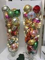 Two large clear glass vases filled with assorted vintage Christmas ornaments in multiple colors and shapes, with string lights inside. Ornaments show vintage style with glitter and painted details.
