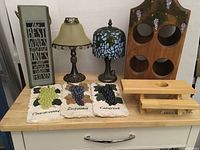 Wine-themed home decor items on wooden table including electric Tiffany-style lamp, vintage lamp, wine bottle holder, decorative plaques and sign box.