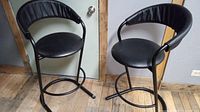 Two vintage Amisco bar stools side by side showing tubular metal frames and black vinyl padded seats and backrests.