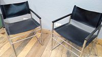 Pair of vintage director chairs with chrome frames and black vinyl seats and backs on wooden floor, showing front and side angles.