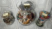 Three large glass jars filled with assorted costume jewelry including necklaces, bracelets, rings, and pins in gold plated and silver colored metals with faux stones