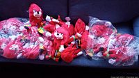Photo showing multiple red Knuckles plush toys with two bags containing approximately 10 plush toys each, displayed on a dark surface.