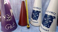 Four vintage mid-century cheerleader horns, including two white with 'Alliance Bulldogs' and bulldog logo, one purple with panther and 'Pat', and one solid brown horn. Measuring tape shows height approximately 25-26 inches.