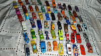 Multiple rows of approximately 60 Hot Wheels and Matchbox diecast toy cars arranged on a fabric surface showing various colors and models.
