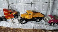 Perspective showing four vintage metal toy trucks arranged in a row on a lace cloth, including a red semi truck, yellow delivery flatbed, pink Tonka truck, and small silver car.