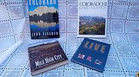 Five large coffee table books laid out showing front covers including two Colorado photo books, Mile High City, Live, and Evergreen High School yearbook.