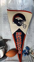 Denver Broncos wool pennant with vintage logo and Broncos lettering hanging against lace curtain, includes small blue Beanie Baby nearby